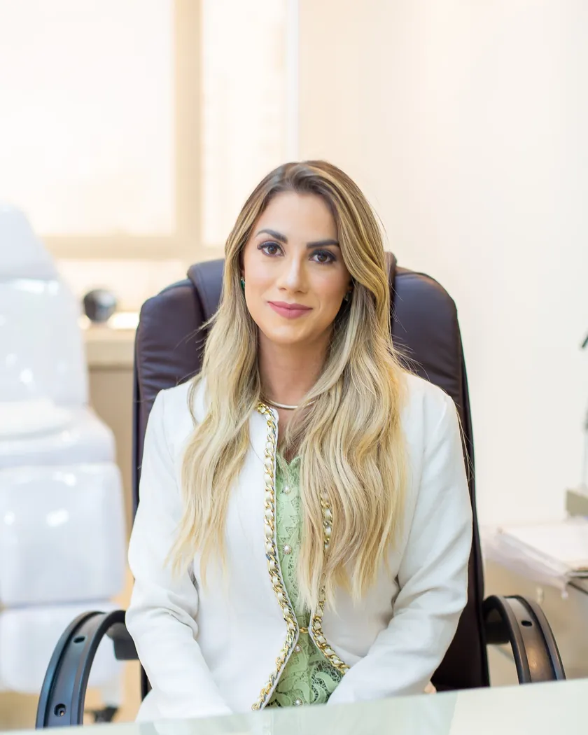 Dra. Alessandra Consone em seu consultório, sentada à mesa, de blazer branco